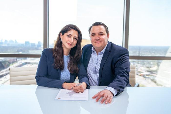 Couple poses for photo in front of glass walls with 达拉斯 cityscape outside. 他们面前的桌子上放着他们签署的礼物协议.